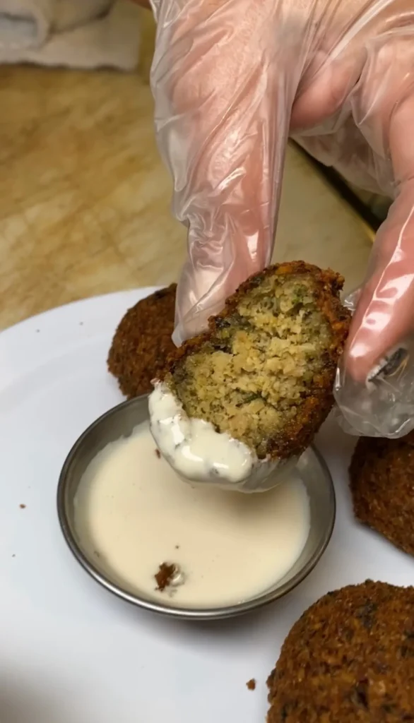 Golden brown falafel ball being dipped into creamy white tahini sauce with gloved hands