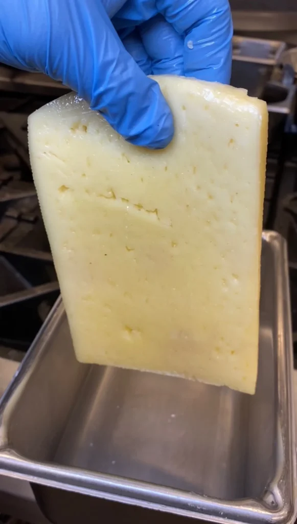 Block of Greek saganaki cheese being held with blue gloves over stainless steel pan in kitchen