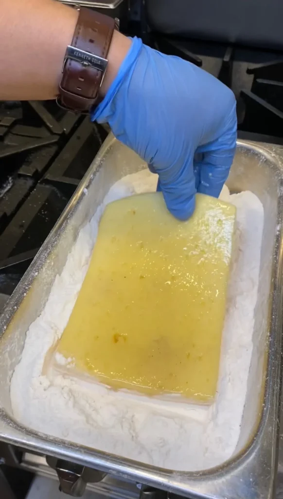 Traditional Greek saganaki cheese melting in a metal pan, being prepared with blue gloves in kitchen