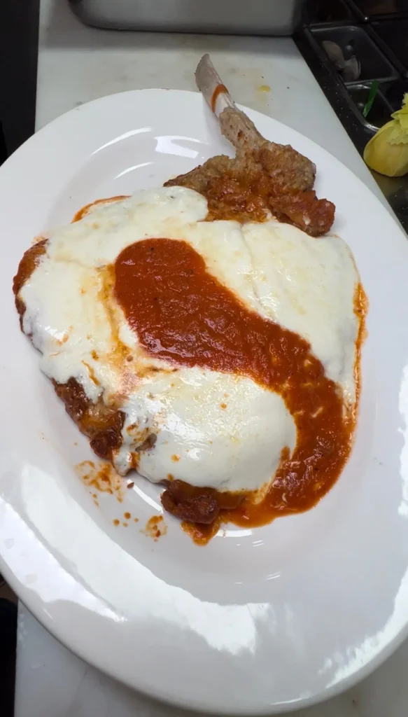 Bone-in thin veal chop parmigiana with melted cheese and tomato sauce on white plate