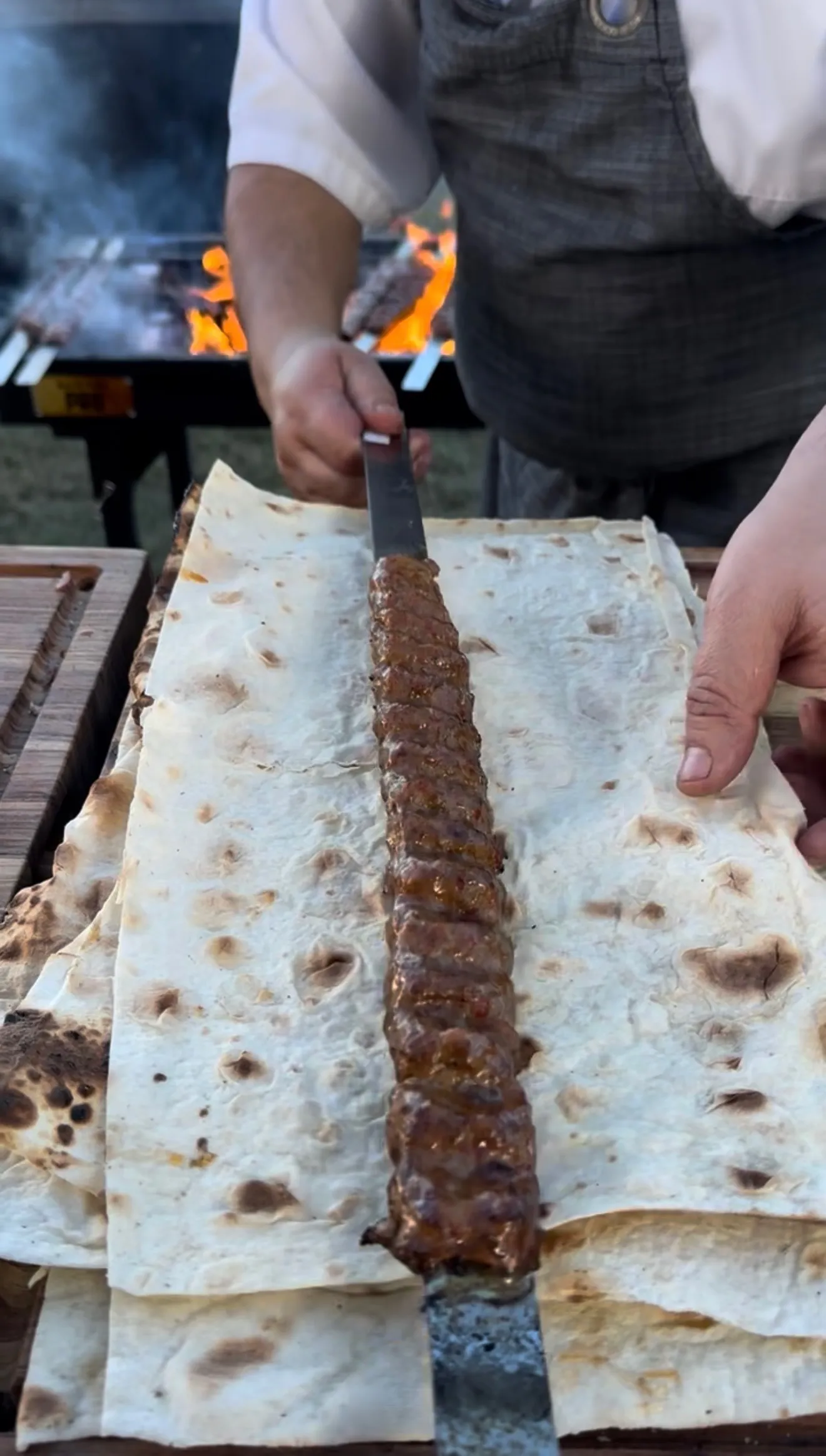 Grilled Adana kebab being sliced with a knife on lavash flatbread over an open flame