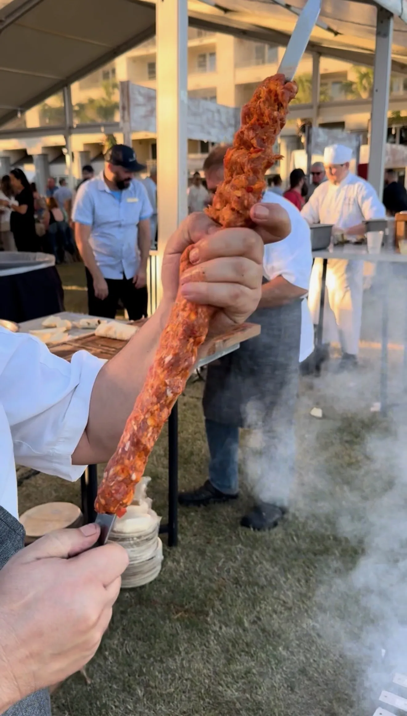Grilled Adana kebab on metal skewer being held over smoky outdoor grill at cooking event