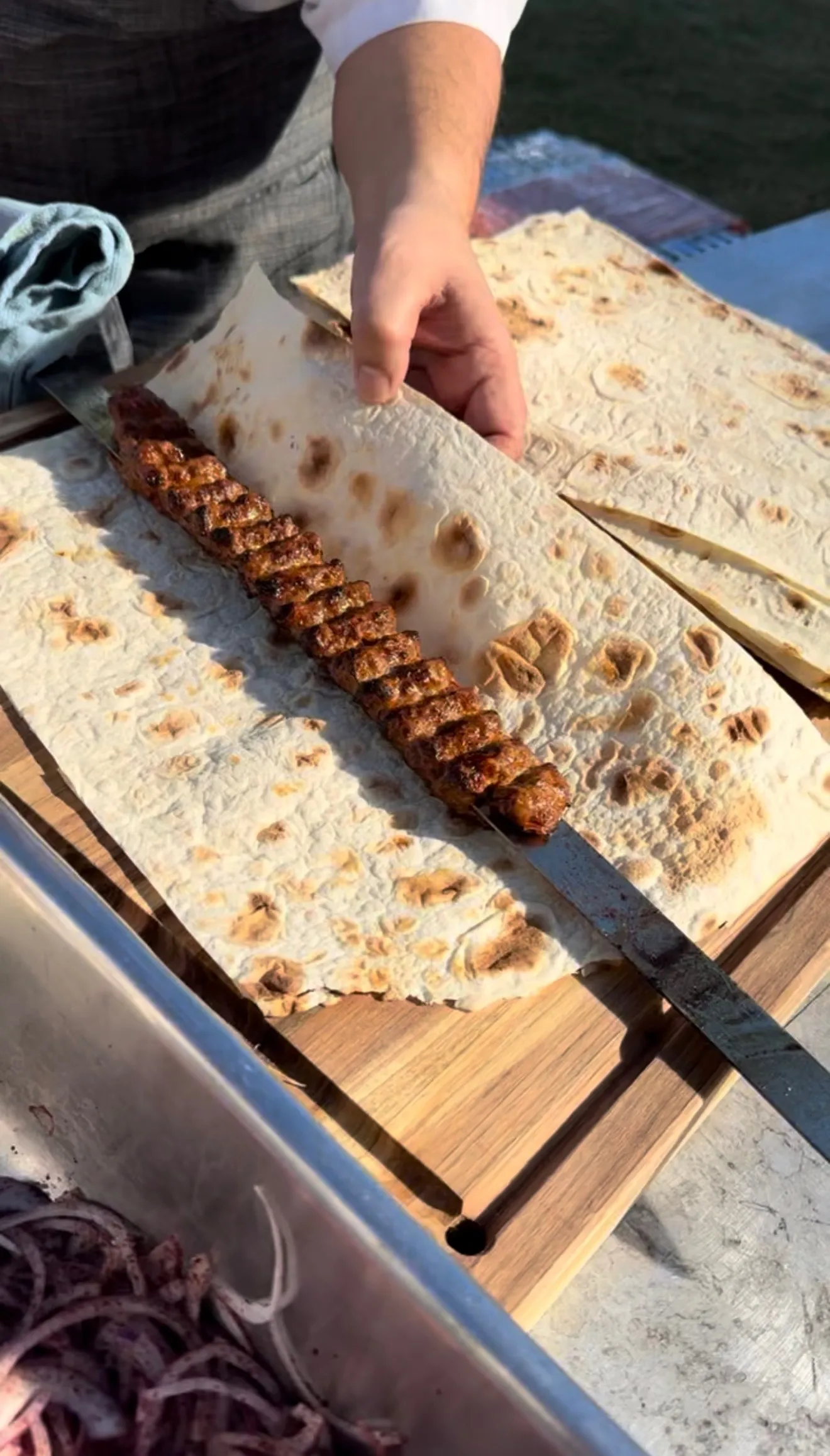Sliced Adana kebab on flatbread with a hand reaching to serve on wooden cutting board
