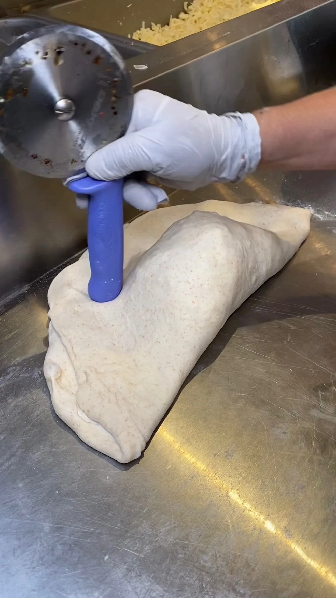 Person in gloves using blue rolling pin to roll out calzone dough on stainless steel kitchen counter