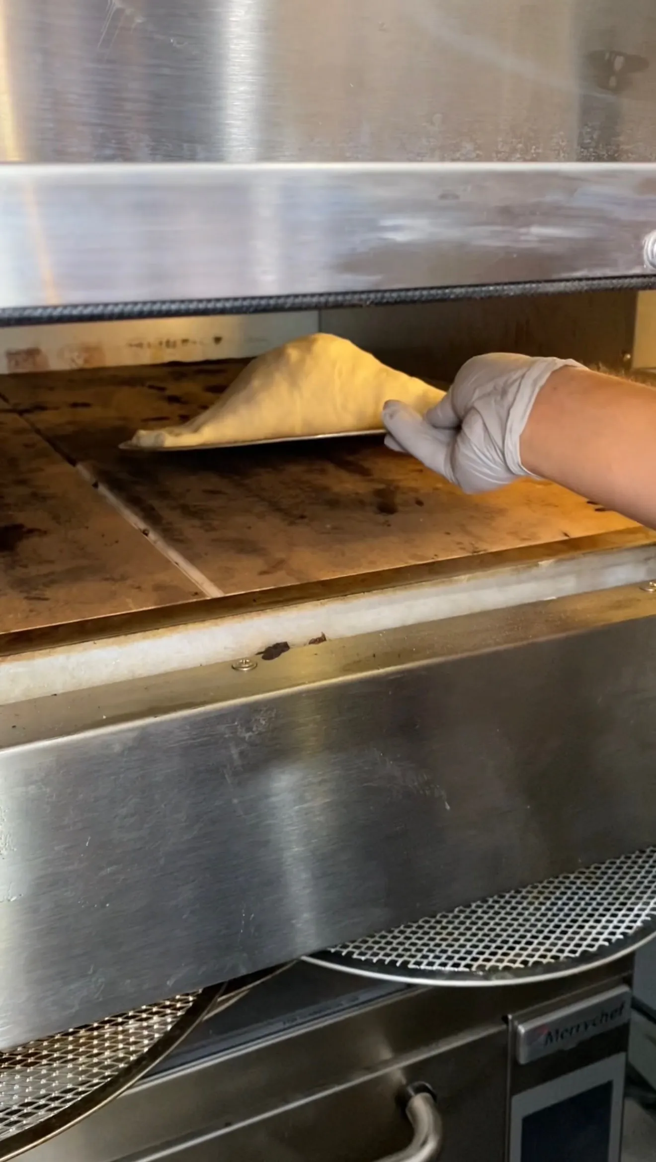 Hand in glove placing raw calzone on wooden peel into commercial pizza oven