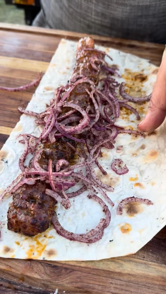 Grilled Adana kebab with charred purple onions on parchment paper over wooden cutting board