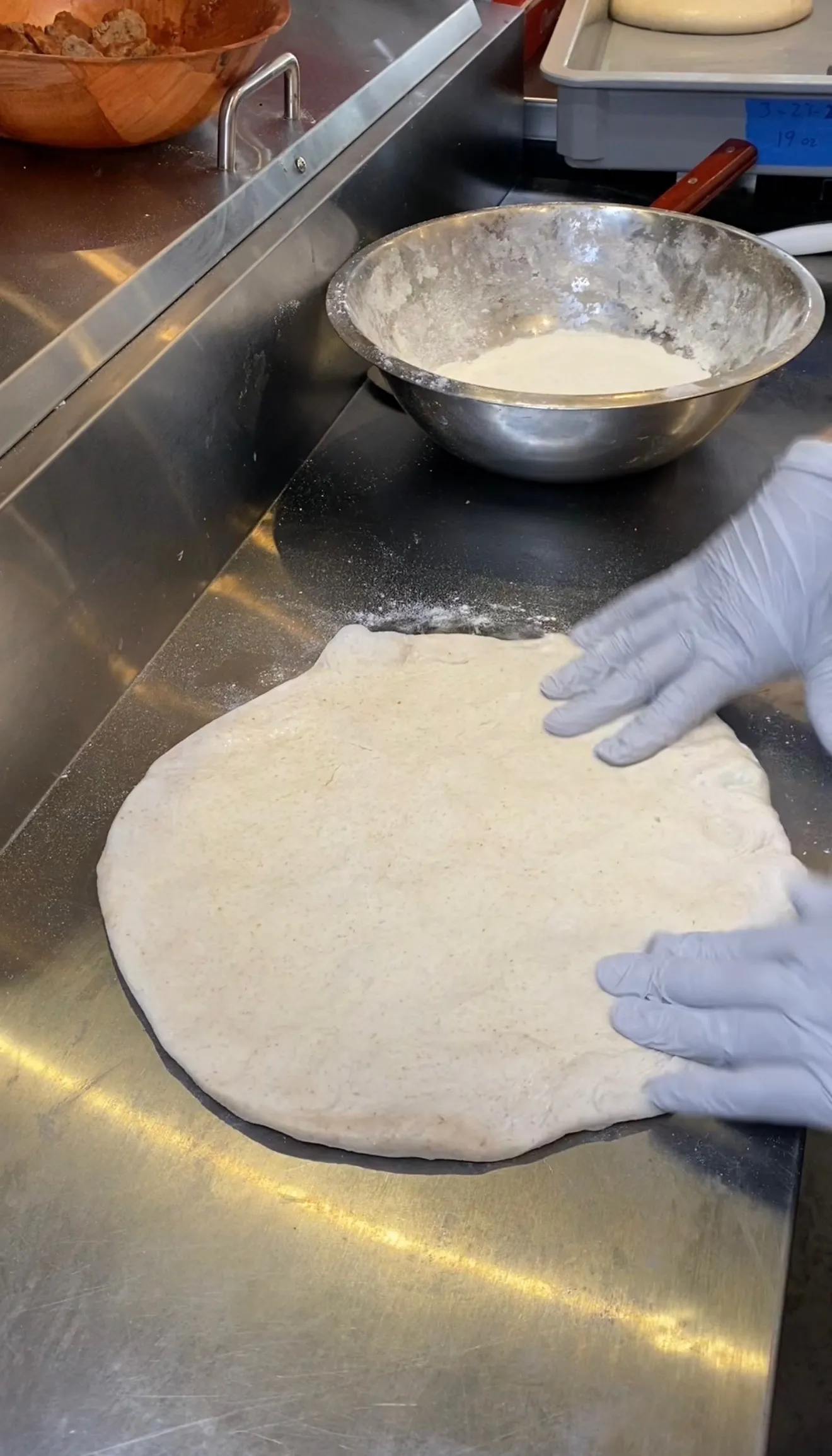 Hands in gloves rolling out pizza dough on stainless steel counter for calzone